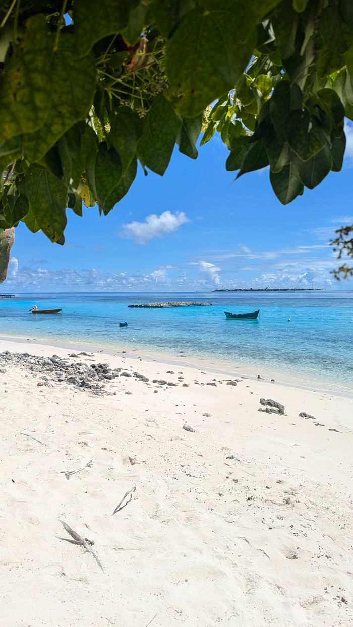 gallery-4 A tranquil beach scene with boats, clear blue sea, and lush greenery under a sunny sky.
